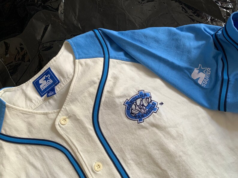 May include: White baseball jersey with blue and black trim. The jersey has a blue and white Starter logo on the right sleeve and a Carolina Tar Heels logo on the front.