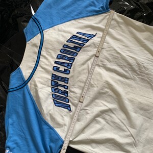 May include: A white and light blue baseball jersey with the text "NORTH CAROLINA" embroidered in blue on the front. The jersey has a blue and white color block design and a star on the left sleeve.