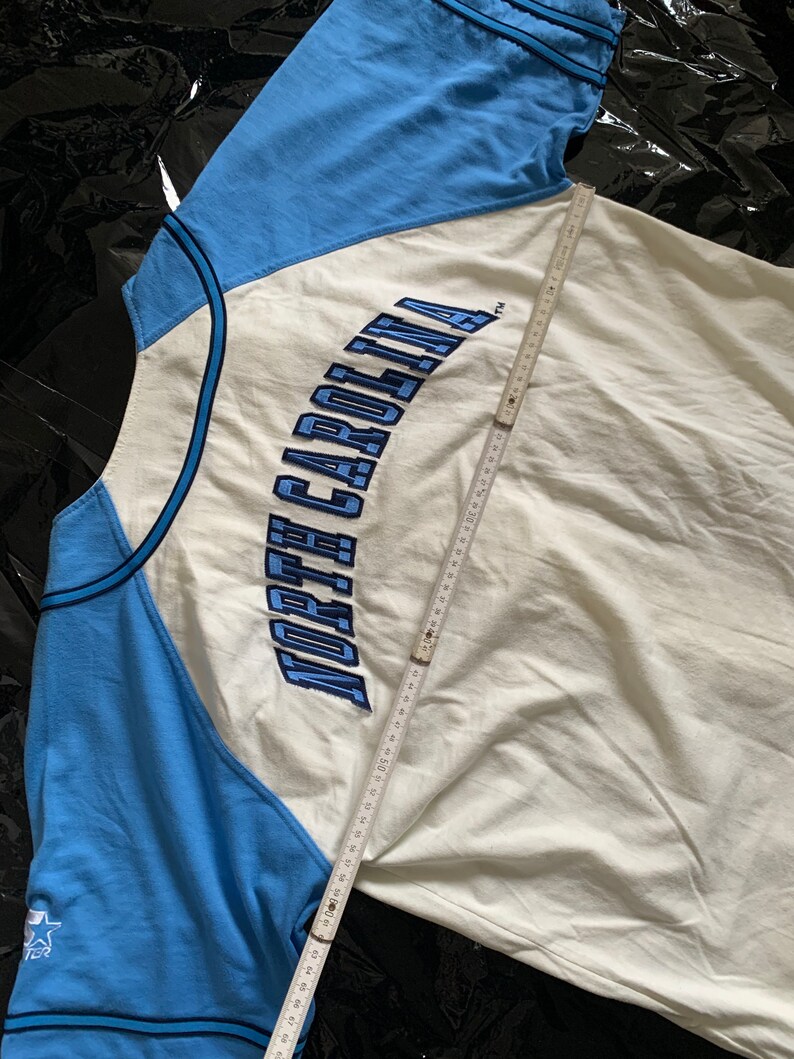 May include: A white and light blue baseball jersey with the text "NORTH CAROLINA" embroidered in blue on the front. The jersey has a blue and white color block design and a star on the left sleeve.