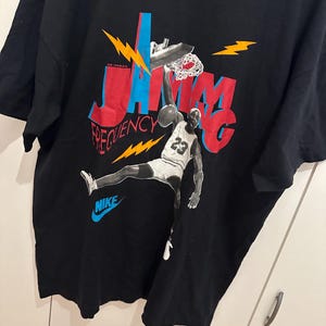 May include: Black t-shirt with a basketball player graphic dunking a ball. The design features the words "Jam Frequency" in red and blue, with lightning bolt accents. The Nike logo is in blue.