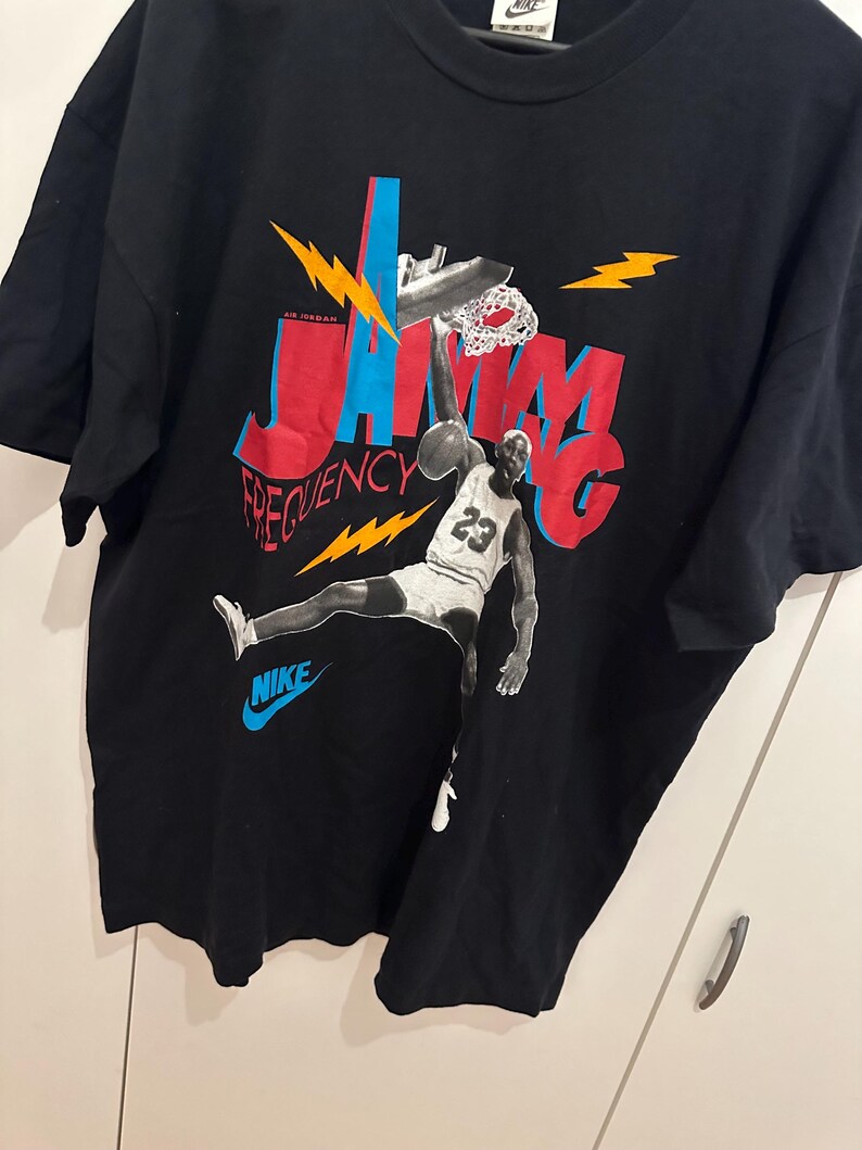 May include: Black t-shirt with a basketball player graphic dunking a ball. The design features the words "Jam Frequency" in red and blue, with lightning bolt accents. The Nike logo is in blue.