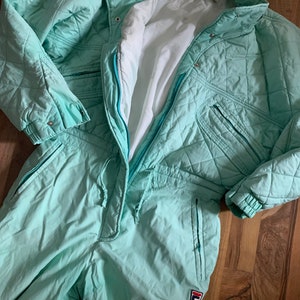 May include: A mint green ski suit with a zipper closure and a Fila logo on the left leg. The suit has a quilted design and multiple pockets.