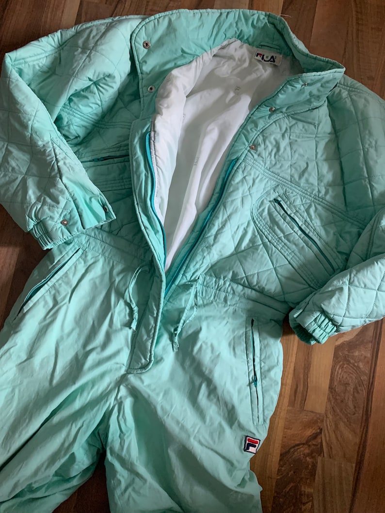 May include: A mint green ski suit with a zipper closure and a Fila logo on the left leg. The suit has a quilted design and multiple pockets.
