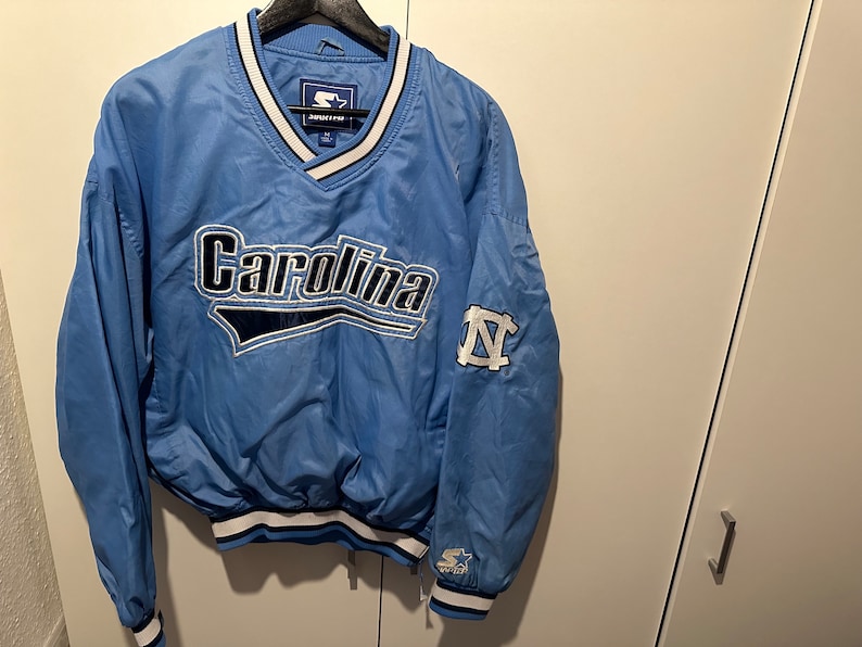K&ouml;nnte beinhalten: Eine hellblaue Vintage-Starterjacke mit einem Carolina-Logo und einem UNC Tar Heels-Logo am &Auml;rmel.