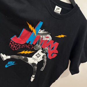 May include: Black Nike t-shirt with a graphic of Michael Jordan in action. The design features the words "JAM" and "FREQUENCY" in red and blue, with yellow lightning bolts. The Nike logo is displayed in blue.