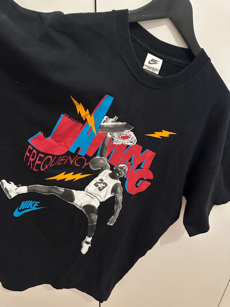 May include: Black Nike t-shirt with a graphic of Michael Jordan in action. The design features the words "JAM" and "FREQUENCY" in red and blue, with yellow lightning bolts. The Nike logo is displayed in blue.