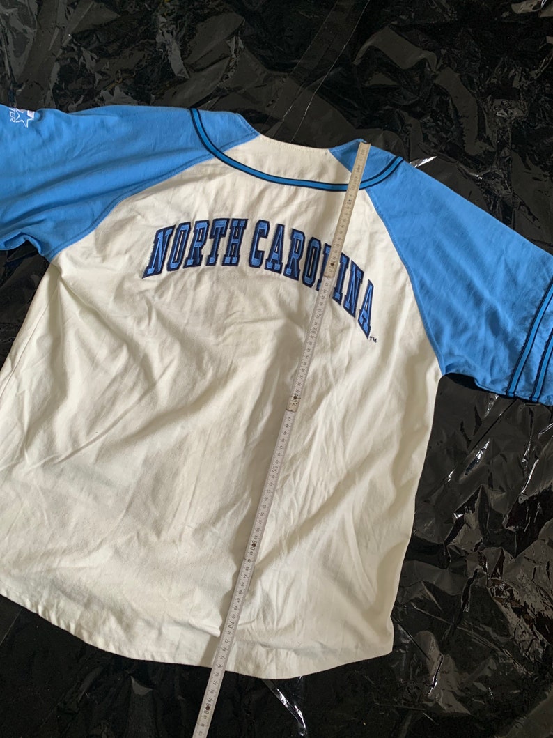 May include: White and light blue baseball jersey with the text "NORTH CAROLINA" in navy blue lettering on the back.