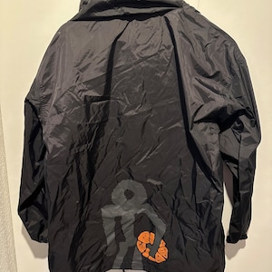 Op de afbeelding: Zwarte capuchontrui met een grote oranje en grijze ankerprint op de rug.