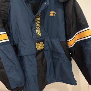May include: A navy blue and black Starter jacket with yellow accents. The jacket features the word "MICHIGAN" in yellow block letters and a yellow "M" logo. The jacket has a hood and a front pocket.