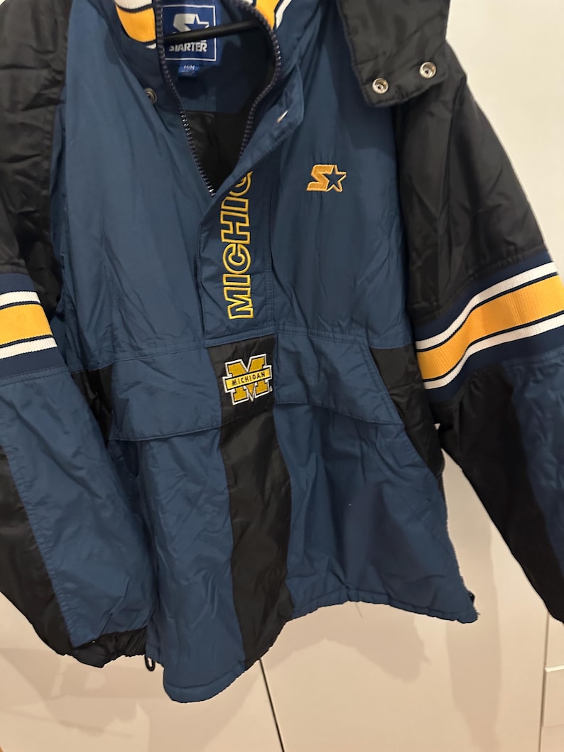 May include: A navy blue and black Starter jacket with yellow accents. The jacket features the word "MICHIGAN" in yellow block letters and a yellow "M" logo. The jacket has a hood and a front pocket.