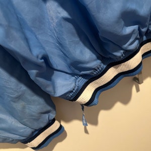 K&ouml;nnte beinhalten: Eine hellblaue Jacke mit wei&szlig;en und dunkelblauen Streifen an den &Auml;rmelb&uuml;ndchen und am Saum. Die Jacke ist aus leichtem Material und hat einen Rei&szlig;verschluss.