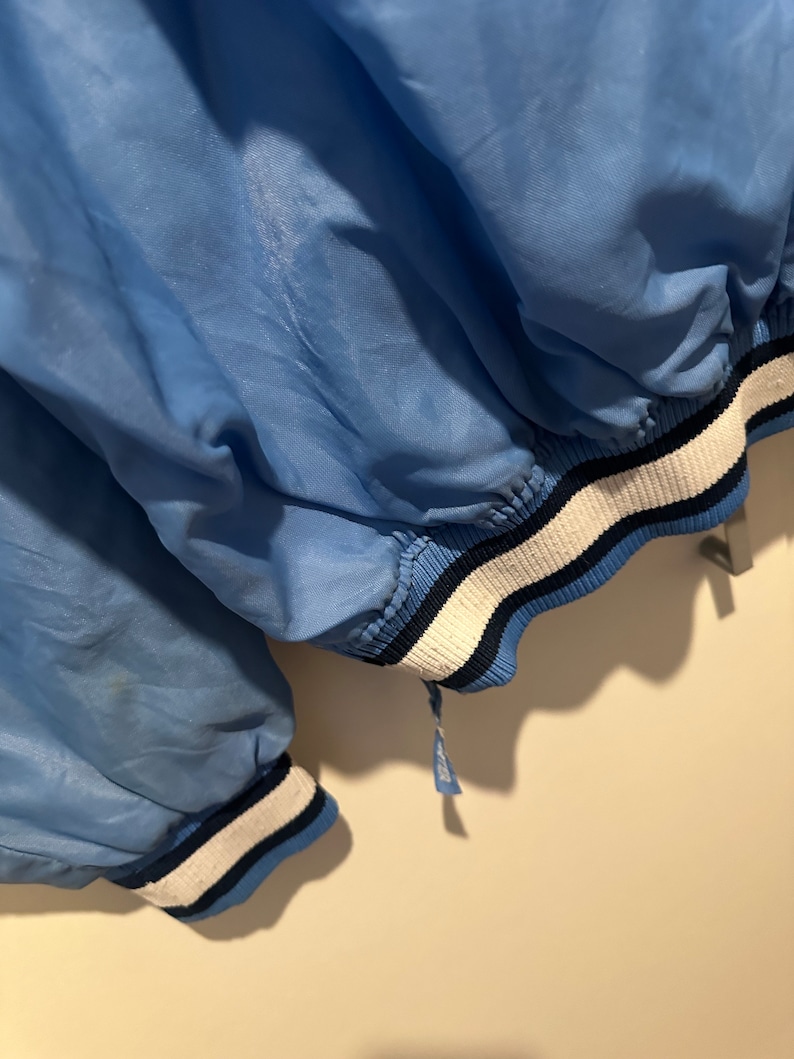 K&ouml;nnte beinhalten: Eine hellblaue Jacke mit wei&szlig;en und dunkelblauen Streifen an den &Auml;rmelb&uuml;ndchen und am Saum. Die Jacke ist aus leichtem Material und hat einen Rei&szlig;verschluss.