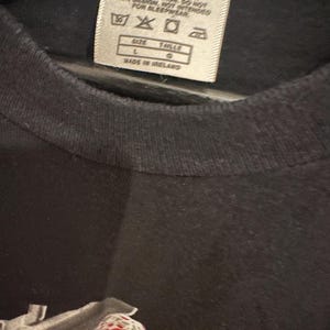 May include: Close-up of a black t-shirt with a tag. The tag includes washing instructions and the text "SIZE L TAILLE G MADE IN IRELAND". The shirt has a graphic design at the bottom.