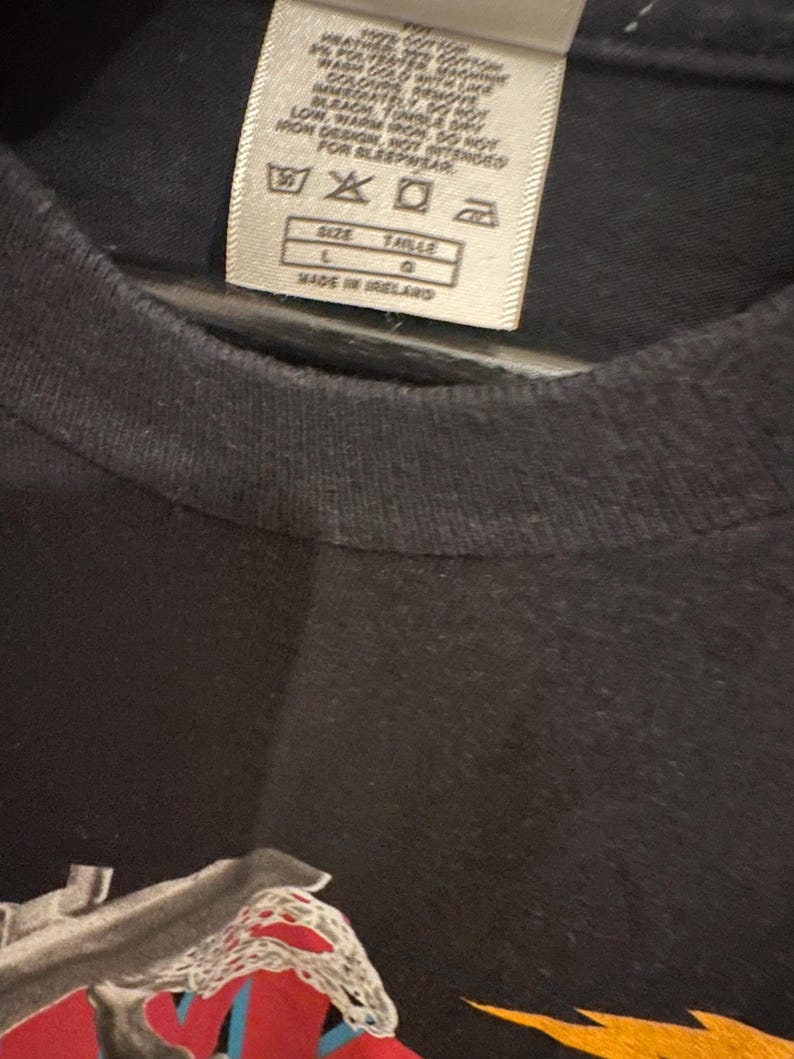 May include: Close-up of a black t-shirt with a tag. The tag includes washing instructions and the text "SIZE L TAILLE G MADE IN IRELAND". The shirt has a graphic design at the bottom.