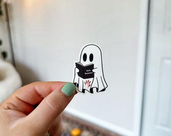 Peut inclure: Un autocollant de fantôme blanc tenant un livre noir qui dit "Dark Romance" avec des empreintes de mains rouges sur la couverture.
