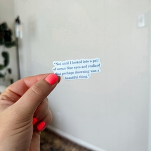 May include: A white sticker with a blue quote that reads "Not until I looked into a pair of ocean blue eyes and realized that perhaps drowning was a beautiful thing."