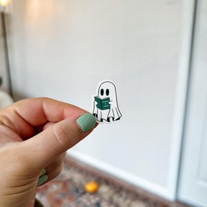 May include: A white ghost sticker with a green book that says "Reading is Fundamental" in a hand holding the sticker.
