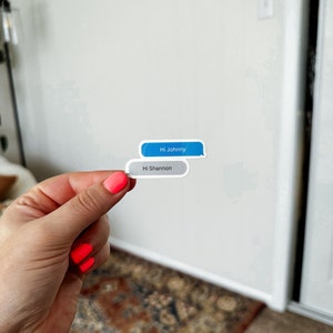 May include: Two stickers in the shape of text message bubbles are held up. The top bubble is blue with the text "Hi Johnny". The bottom bubble is gray with the text "Hi Shannon".