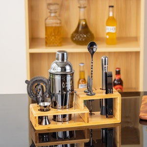 May include: A stainless steel cocktail shaker set with a bamboo organizer. The set includes a shaker, strainer, jigger, bar spoon, and other tools. The tools are black and silver, and the set is displayed on a black surface.