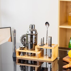 May include: A stainless steel cocktail shaker set with a wooden organizer. The set includes a shaker, jigger, muddler, strainer, bar spoon, and other tools. The set is displayed on a black surface.