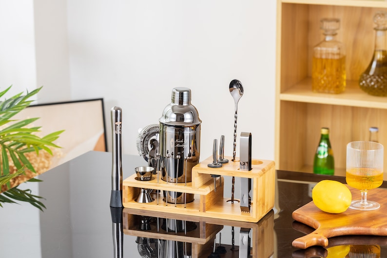 May include: A stainless steel cocktail shaker set with a wooden organizer. The set includes a shaker, jigger, muddler, strainer, bar spoon, and other tools. The set is displayed on a black surface.