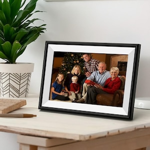 May include: A black framed digital photo frame displaying a family portrait. The frame is on a wooden surface next to a potted plant. The family is gathered in front of a Christmas tree.