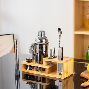 May include: A stainless steel cocktail shaker set with a wooden stand. The set includes a shaker, strainer, jiggers, muddler, bar spoon, and other bar tools. A lemon and a glass of liquid are on a wooden cutting board.