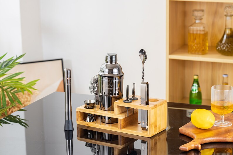 May include: A stainless steel cocktail shaker set with a wooden stand. The set includes a shaker, strainer, jiggers, muddler, bar spoon, and other bar tools. A lemon and a glass of liquid are on a wooden cutting board.
