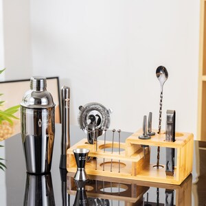 May include: A stainless steel cocktail shaker and bar tools set. The set includes a shaker, strainer, jigger, muddler, bar spoon, and other tools, all displayed in a wooden organizer. The tools are arranged on a black surface.