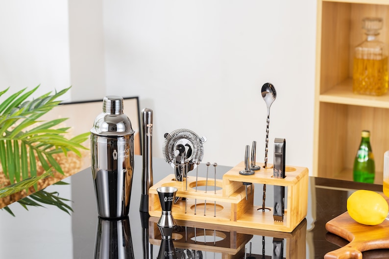 May include: A stainless steel cocktail shaker and bar tools set. The set includes a shaker, strainer, jigger, muddler, bar spoon, and other tools, all displayed in a wooden organizer. The tools are arranged on a black surface.