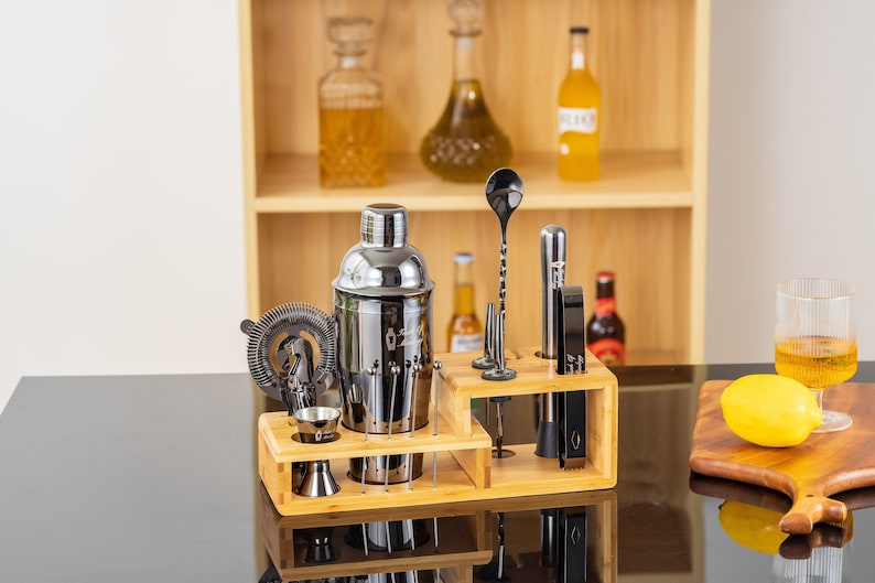 May include: A stainless steel cocktail shaker set with a bamboo organizer. The set includes a shaker, strainer, jigger, bar spoon, and other tools. The tools are black and silver, and the set is displayed on a black surface.