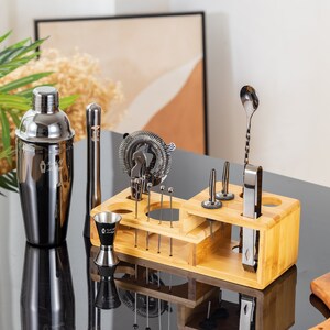 May include: A black and silver cocktail shaker and bar tool set displayed on a black surface. The set includes a shaker, jigger, strainer, muddler, and other tools, all neatly organized in a bamboo holder. A lemon and a glass are also visible.