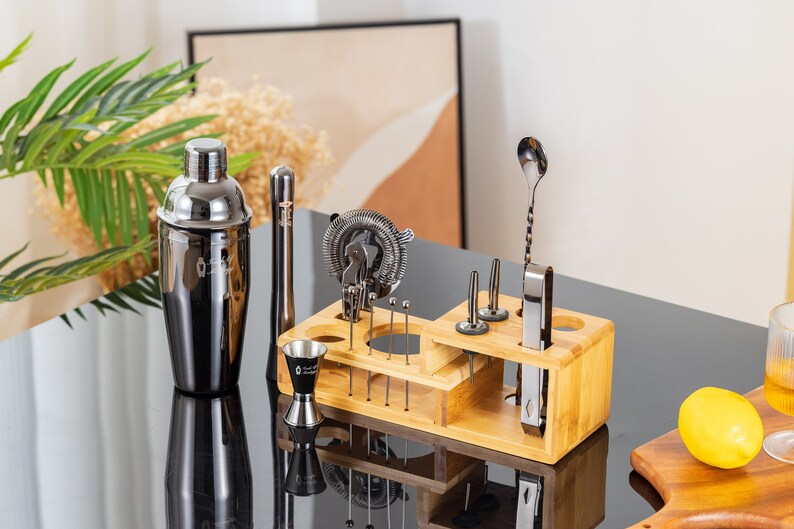 May include: A black and silver cocktail shaker and bar tool set displayed on a black surface. The set includes a shaker, jigger, strainer, muddler, and other tools, all neatly organized in a bamboo holder. A lemon and a glass are also visible.