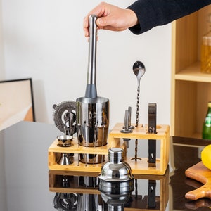 May include: A stainless steel bar tool set with a muddler being held over a shaker. The set includes a strainer, jiggers, a bar spoon, and other tools, all displayed on a wooden stand. A lemon and a glass of liquid are on a wooden cutting board.