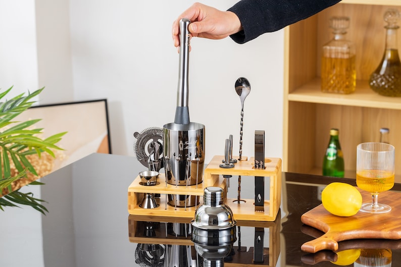 May include: A stainless steel bar tool set with a muddler being held over a shaker. The set includes a strainer, jiggers, a bar spoon, and other tools, all displayed on a wooden stand. A lemon and a glass of liquid are on a wooden cutting board.