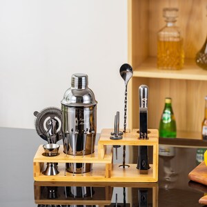 May include: A stainless steel cocktail shaker set with a wooden stand. The set includes a shaker, strainer, jigger, bar spoon, muddler, and other bar tools. The set is displayed on a black surface.