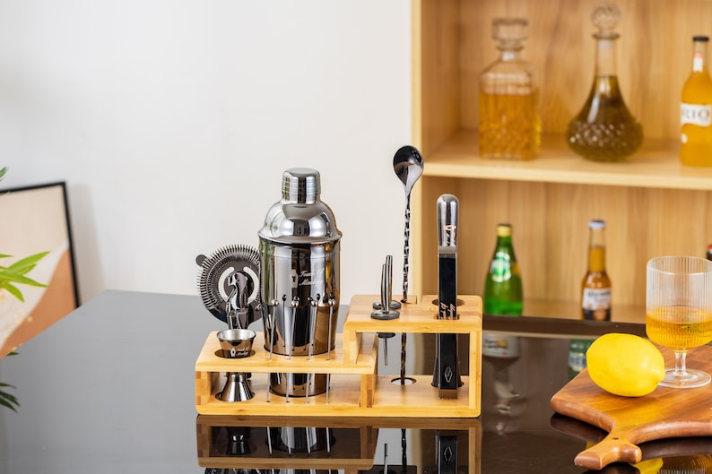 May include: A stainless steel cocktail shaker set with a wooden stand. The set includes a shaker, strainer, jigger, bar spoon, muddler, and other bar tools. The set is displayed on a black surface.