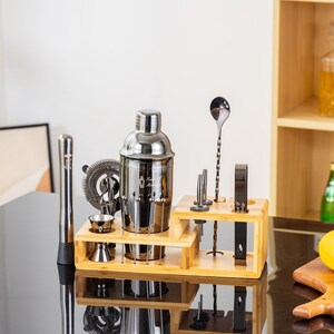 May include: A stainless steel cocktail shaker set with various bar tools, including a muddler, strainer, and jigger, arranged on a bamboo stand. The set is displayed on a reflective black surface, with bottles and a glass of liquid in the background.