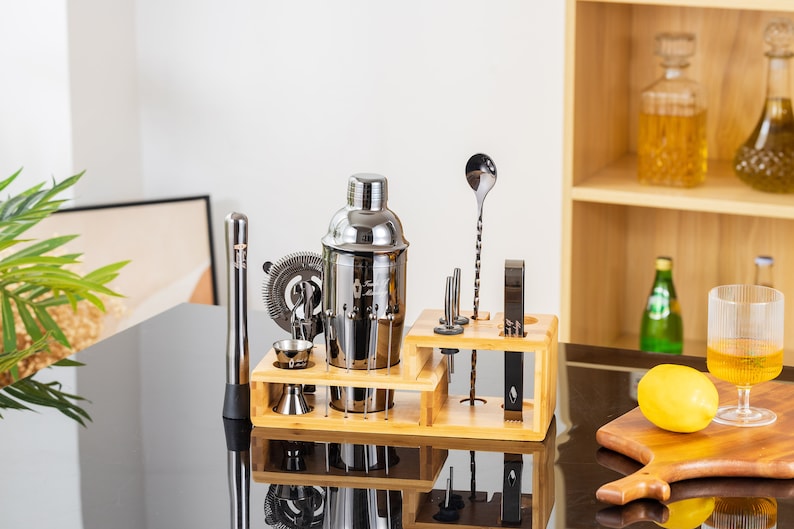 May include: A stainless steel cocktail shaker set with various bar tools, including a muddler, strainer, and jigger, arranged on a bamboo stand. The set is displayed on a reflective black surface, with bottles and a glass of liquid in the background.
