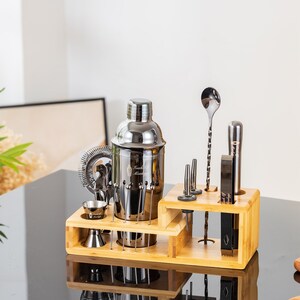 May include: A stainless steel cocktail shaker set with a bamboo wood stand. The set includes a shaker, strainer, jiggers, muddler, bar spoon, and other bar tools. The tools are arranged on a two-tiered bamboo stand.