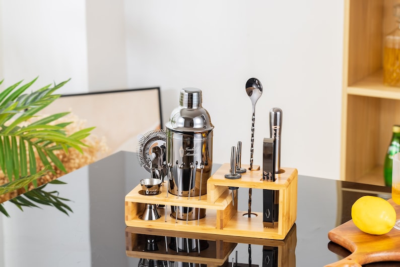 May include: A stainless steel cocktail shaker set with a bamboo wood stand. The set includes a shaker, strainer, jiggers, muddler, bar spoon, and other bar tools. The tools are arranged on a two-tiered bamboo stand.