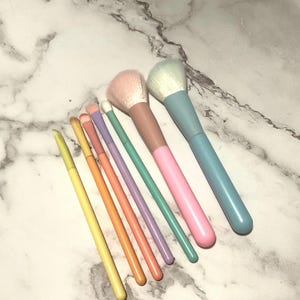 May include: A collection of pastel-colored makeup brushes. The set includes brushes with pink, peach, green, purple, yellow, and orange handles. The brush heads are in shades of pink and white. The brushes are arranged on a white and gray marble surface.