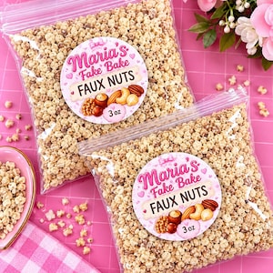 May include: Two clear plastic bags filled with small, beige, textured faux nut pieces. Each bag features a round label with the text Maria's Fake Bake FAUX NUTS 3 oz, decorated with illustrations of various nuts and pink hearts.