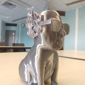 May include: A silver 3D printed figurine of a person wearing sunglasses. The figurine is sitting with its legs crossed and has a detailed face.