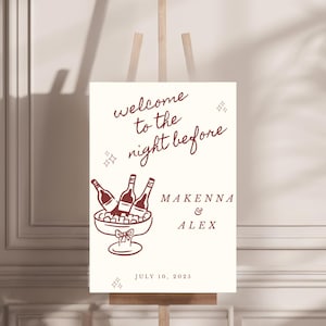 May include: A cream-colored sign on a wooden easel with burgundy text and illustrations. The sign reads "welcome to the night before" with the names "Makenna & Alex" and the date "July 10, 2025". The illustration depicts wine bottles in an ice bucket.