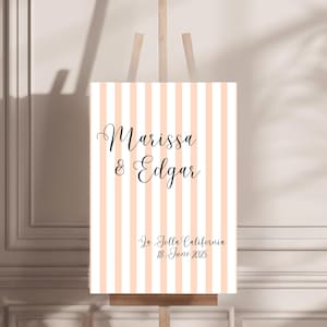 May include: A wedding welcome sign with vertical peach and white stripes. The sign displays the names "Marissa & Edgar" in elegant script, along with the location "In Jolla California" and the date "18 June 2025". The sign is mounted on a wooden easel.