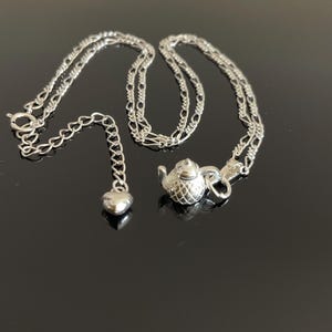 May include: A silver necklace with a teapot charm and a heart charm. The chain is a delicate, silver curb chain.