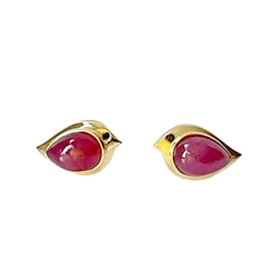 May include: A pair of gold-colored bird-shaped earrings. Each earring features a teardrop-shaped, deep red gemstone set within the bird's body. The bird's head has a small black eye and a yellow beak. The earrings are set against a white background.