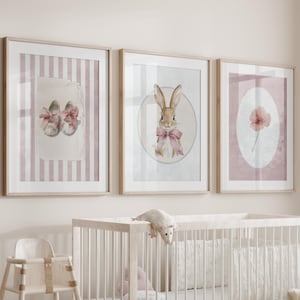 Pode incluir: Um berço branco com um conjunto de roupa de cama estampada em branco e cinza. Três impressões emolduradas com ilustrações rosa e brancas de um coelho, uma flor e um par de sapatos estão penduradas na parede acima do berço. Uma cadeira alta branca está em primeiro plano.