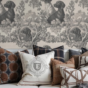 May include: A white sofa with several decorative pillows in shades of brown, cream, and black. The pillows feature various patterns, including geometric designs and a monogram. The background is a wallpaper with a repeating dog and tree design.